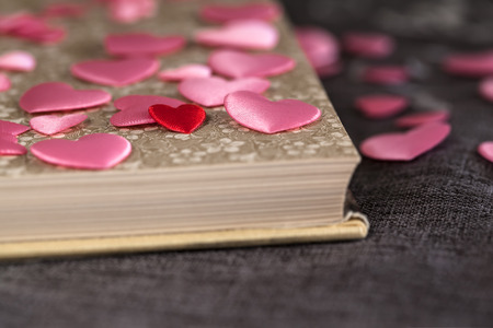 Detail of one very small red heart among many pink ones and open vintage book on brown textile...