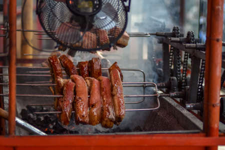 Large cuts of pork belly being roasted over hot charcoals and being turned evenly by use of a motor, cogs and a chain in the Philippines.の写真素材