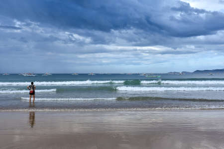 A young woman standing ankle deep in a tropical sea looking out at traditional filipino pump boats and the passing storm clouds.の写真素材