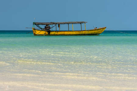 A Old yellow long tail boat anchored just off of shore in a crystal clear tropical sea.の写真素材