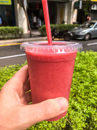 A male human hand holding up a clear plastic cup of a cold Strawberry Smoothie with a red straw and...
