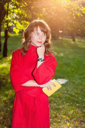 Beautiful woman in red coat posing in autumn Park, keeping in hand little yellow bookの写真素材