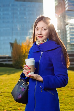 Asian woman holding coffee in hand, standing outdoors behind skyscraper in the sunlight. enjoying her morning coffee in the sunlight. Woman smaling happy in her 20sの写真素材