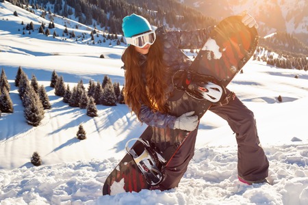 Girl snowboarder stands mountain top with snowboard in hands, at sunset. Mountains in the backgroundの写真素材