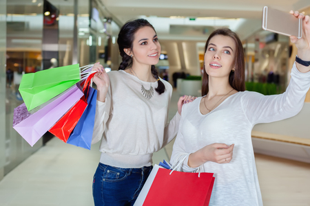 Two beautiful girls makes selfie in a shopping centre with a bag of gifts. Smiling posing for the camera. To make purchasesの写真素材