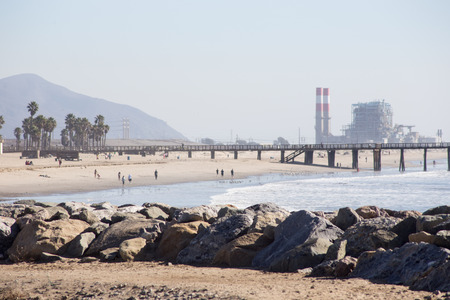 A sunny day in California at the beach  Tourists walk along the shore  Rocks, a pier and power plant...