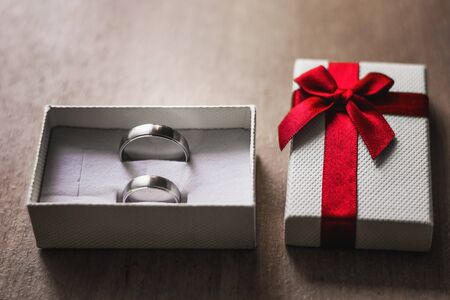 Marry me concept. White box with two engagement rings and red ribbon on a wooden table.の写真素材