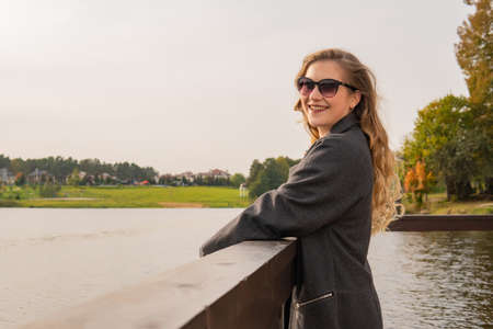 The girl stands near the water and smiles in sunglasses, in her coatの写真素材