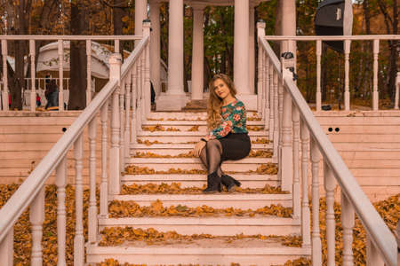 A girl sits on a white staircase in an autumn park in the woods. Beautiful blondの写真素材