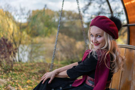 a girl sits on a garden swing, forged in a burgundy coat and biret, with a charismatic appearance,...