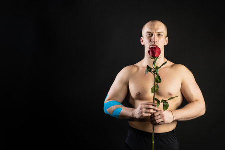 Athletic man holding a red rose, naked for the day of velentin pumped up, on his arm a teip of a bodybuilder romantic. lover guy LGBT. In the studio on a black background Joyful with tender lipsの写真素材