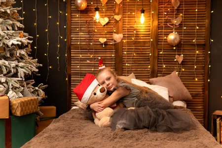 A girl hugs a bear toy in a Christmas cap bear Christmas kid child little, from people home for play sitting room, small joyful. Bedroom bed holding, background wakeの写真素材