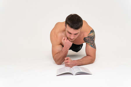 Bodybuilder reads the book on a white background isolated at the bottom of his head on his hands man young reading, athletic bodybuilding muscles, Isolated gym, posing tanの写真素材