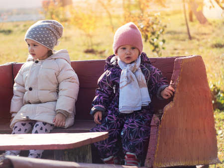 portrait of two children in autumn jackets, taken on a sunny autumn day, sit on a bench in a city parkの写真素材