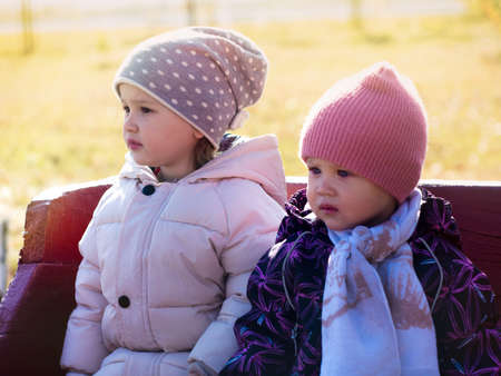 Portrait of two children in autumn jackets, taken on a sunny autumn day, sit on a bench in a city parkの写真素材