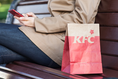 Minsk, Belarus, june 21, 2017: lunch bag KFC on a bench next to a woman with a mobile phone. Kentucky Fried Chicken (KFC) is world's second largest restaurant chain almost 20,000 locations globally.のeditorial素材