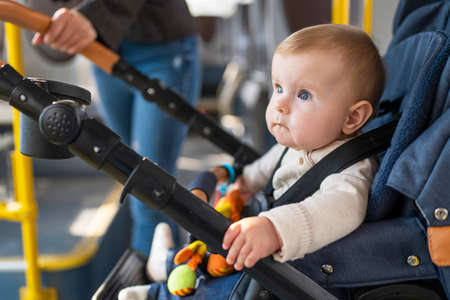 A cute little baby of 10 months is sitting in a stroller. A child is fastened with seat belts in stroller rides a passenger bus with her mother. Concept of child safety.の写真素材