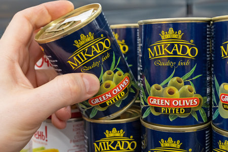 Cans of canned green olives brand Mikado. A customer takes a can of green olives from a supermarket shelf. Minsk, Belarus 2022のeditorial素材