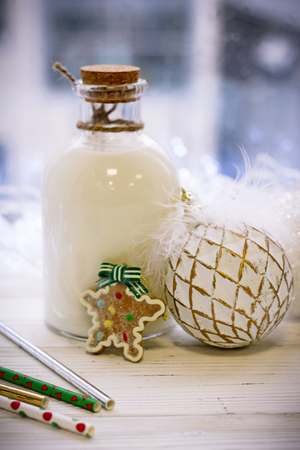 New Year's composition, a bottle of milk or cream on a wooden table, Christmas toys: balls, lights, sticks, and sweet biscuits and gingerbread on a sunny dayの写真素材