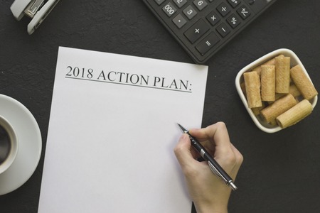 Hand writind 2018 action plan, a white bowl of cookies, coffee and office supplies on a black concrete background, top viewの写真素材