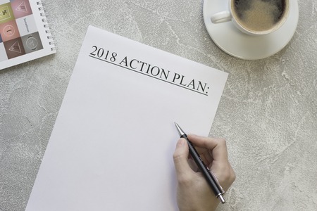 Hand writing 2018 action plan, a cofee cup with saucer, a notebook on a light concrete background,...