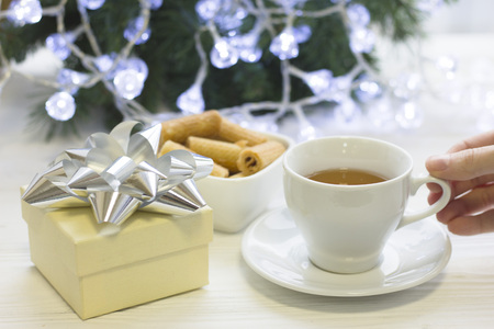 A hand holding a cup of tea, a bowl of cookies, a gift box, Christmas tree on the backの写真素材