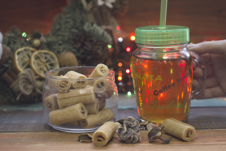 Hand holding a glass clear cup of tea with a cap, a bowl of cookies, a Christmas wreathの写真素材