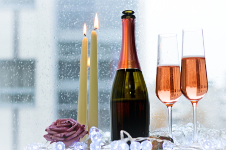 A festive morning for a couple. Two filled glasses and a bottle of rose sparkling wine with two burning candles. A lilac rose and white fairy lights on a white wooden table. Rainy day. Light backgroundの写真素材