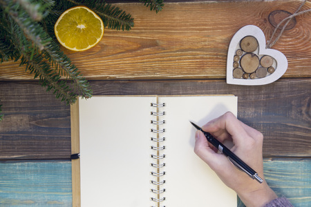 A colored wooden Christmas or New Year background with fir tree branches, a hand writing in a blank notebook and a wooden toy heartの写真素材