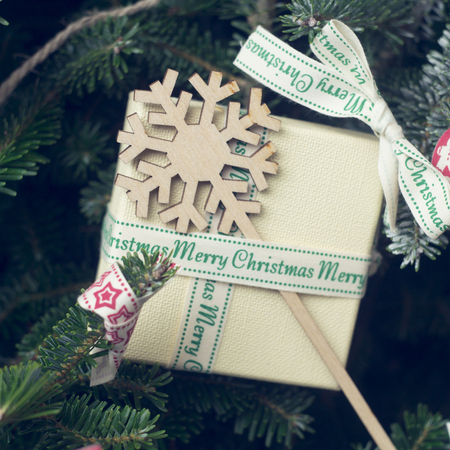 Cristmas gift box among fir tree branches. A beige gift box tied with ribbon with Merry Christmas inscription and a flat wooden snowflake. Close upの写真素材