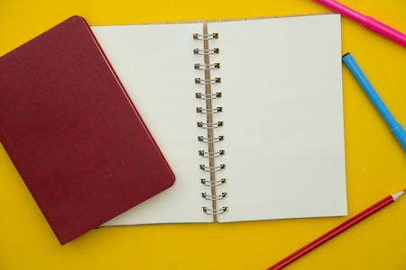 Write down creative ideas. An opened blank notebook, a closed notebook in a red cover and colored markers and pencils on a yellow background. Space for your text or product display.の写真素材