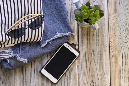 Preparation for travel concept- small plant, patterned summer clothes, denim jeans, aviator sunglasses and cell phone on a wooden background. Top view.の写真素材