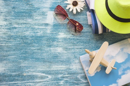 Traveler essentials on blue wooden background- a wooden toy airplane, a notebook, sunglasses and a bright green hat. Space for a text or product display, top view.の写真素材