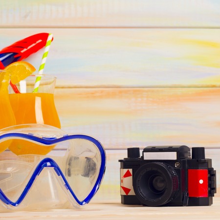 Enjoy a taste of fresh fruit at the beach. A vintage camera, two glasses of orange juice with colorful straws and a diving mask. Close up, square crop.Light colored wooden background. Travel and vacation concept.の写真素材
