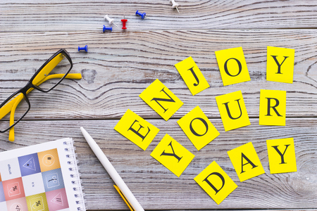 Enjoy your day printed inscription, glasses, a pen, a notebook and pins on a light wooden table, top view.の写真素材