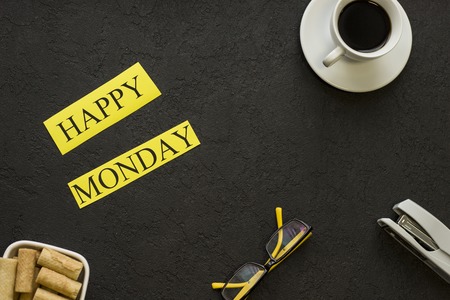 Happy monday inscription with a cup of hot coffee, a bowl of cookies and office items. Black...