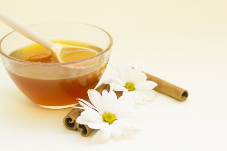 Beauty and skin care natural ingredients- a bowl of fresh honey with a wooden spoon, camomile flowers and cinnamon sticks on a light background. Close up. Spa and body care concept.の写真素材