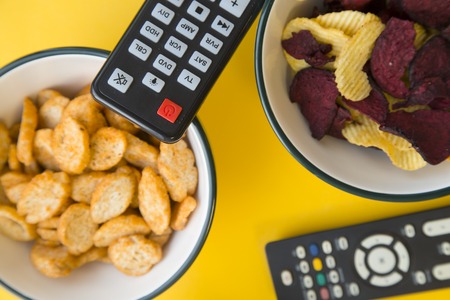 Weekend at home with a movie. Two bowls with salty croutons and potato and beetrrot,chips a pair of glasses and two remote controls on a bright one-color yellow background. close up.の写真素材