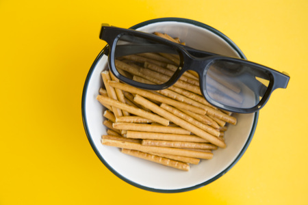Weekend at home with tv shows. A pair of glasses on a bowl with delicious salty bread sticks on a bright one-color yellow background. Close up. Space for a text or product displayの写真素材