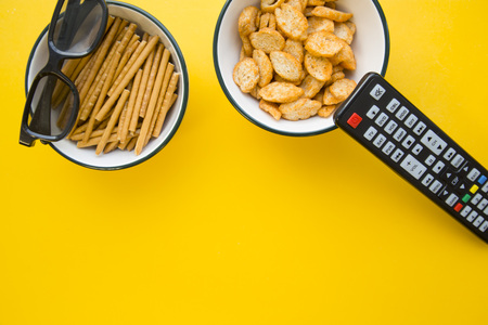 Weekend at home with tv shows. Glasses on a bowl with salty bread sticks, a bowl of fresh croutons and a remote control, bright one-color yellow background. Space for a text or product displayの写真素材