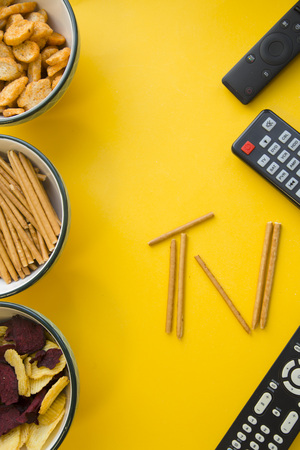 Weekend, leisure and hobby concept. A TV inscription made of bread sticks in a vertical frame of snacks in bowls and remote controls on a bright one-colore yellow background.の写真素材