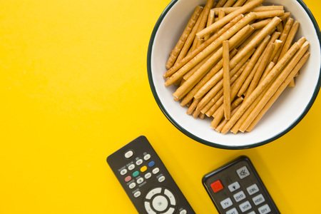 Watching tv programms at home. Weekend, hobby and leisure concept. A bowl of fresh salty bread sticks and two remote controls on a bright one-color yellow background. Close upの写真素材