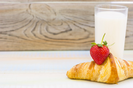 Tasty Breakfast Concept with a glass of milk and a buttery croissant with a strawberry on a light wooden table against rustic wooden wallの写真素材