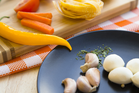 Preparing Healthy Food, Concept. Ingridients for cooking pasta with, carrot, tomato and chili pepper on a wooden cutting board and orange plaid cloth, mozzarella and garlic on a black plateの写真素材