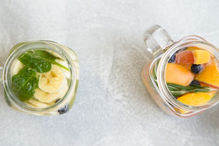 Two glass jars of detox infused water with sliced babanas, grapefruit, blueberry, rosemary and spinach, light concrete background, top viewの写真素材