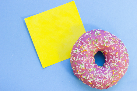 A pink glazed donut and an empty yellow sticker note on a blue background with copy spce, top view