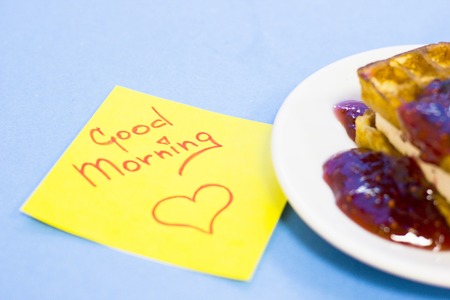 Delicious breakfast: a plate of fresh waffles with raspberry jam and Good Morning yellow sticker note on a blue background with copy spaceの写真素材