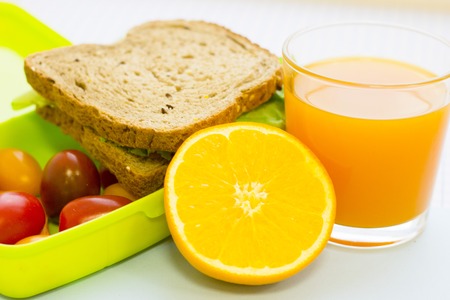 A green plastic lunch box with fresh vegetables,rye bread sandwich, orange and a glass of juice, close up, white squared paper backgroundの写真素材