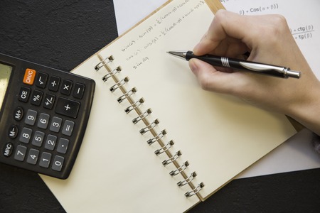 Woman's hand writing trigonometry formulas in a notebook with a pen and calculator, balck stone...