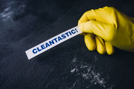 Cleaning office or house concept. Mens hand in yellow rubber glove holding Cleantastic inscription sign, concrete dark background, top view, copy spaceの写真素材